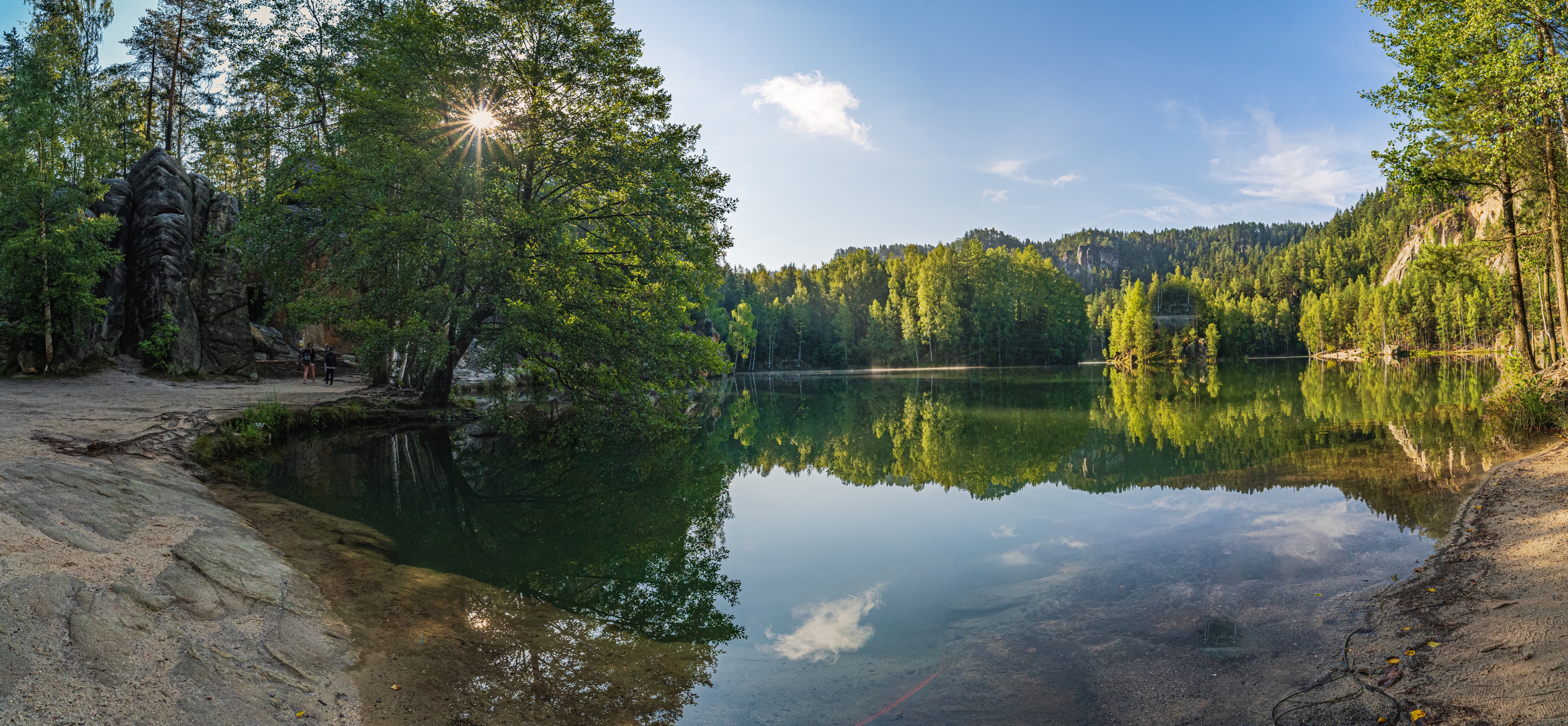 Un étang rocheux à Adršpach, entouré de forêts et de rochers, avec un reflet parfait des arbres et du ciel dans l'eau et un rayon de soleil qui brille à travers les feuilles.