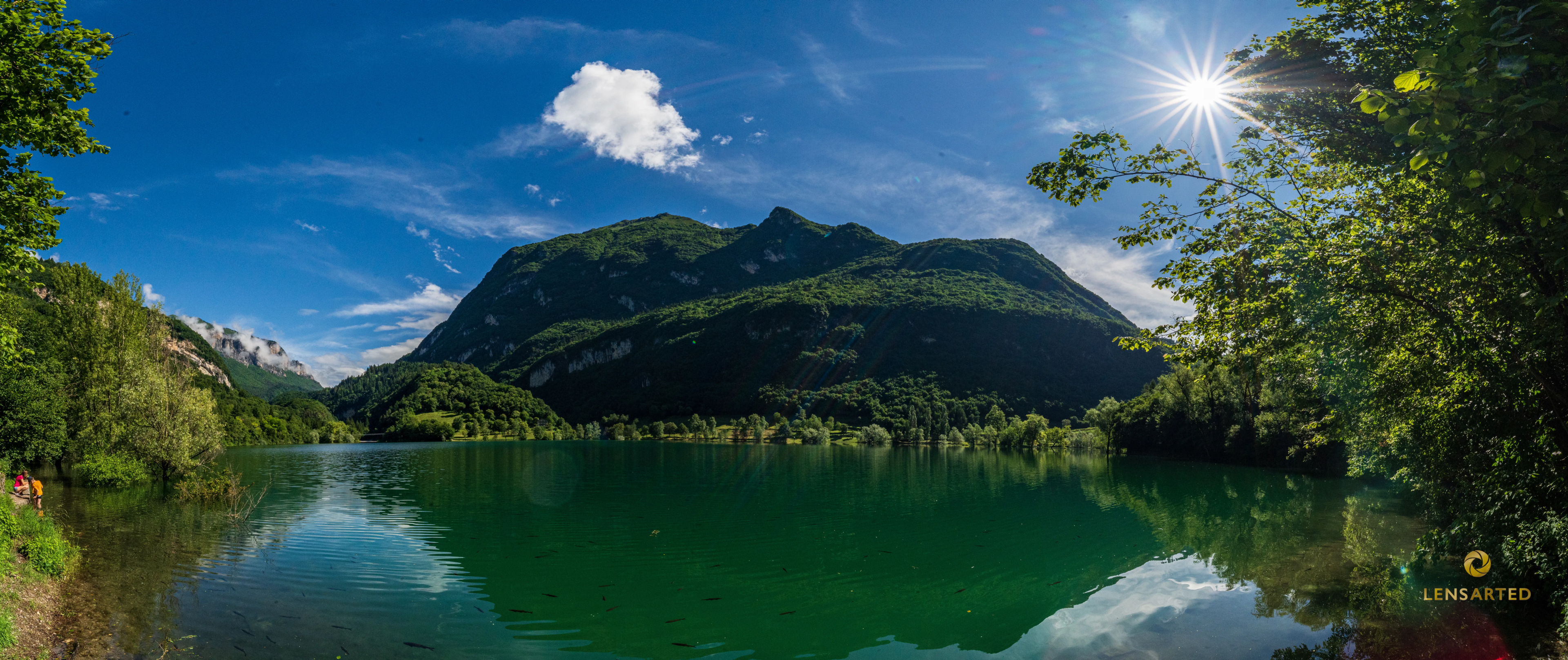 Le lac Tenno, entouré de montagnes et de forêts, avec le ciel qui se reflète dans ses eaux calmes et turquoise, et un rayon de soleil qui perce à travers les branches sur la droite.