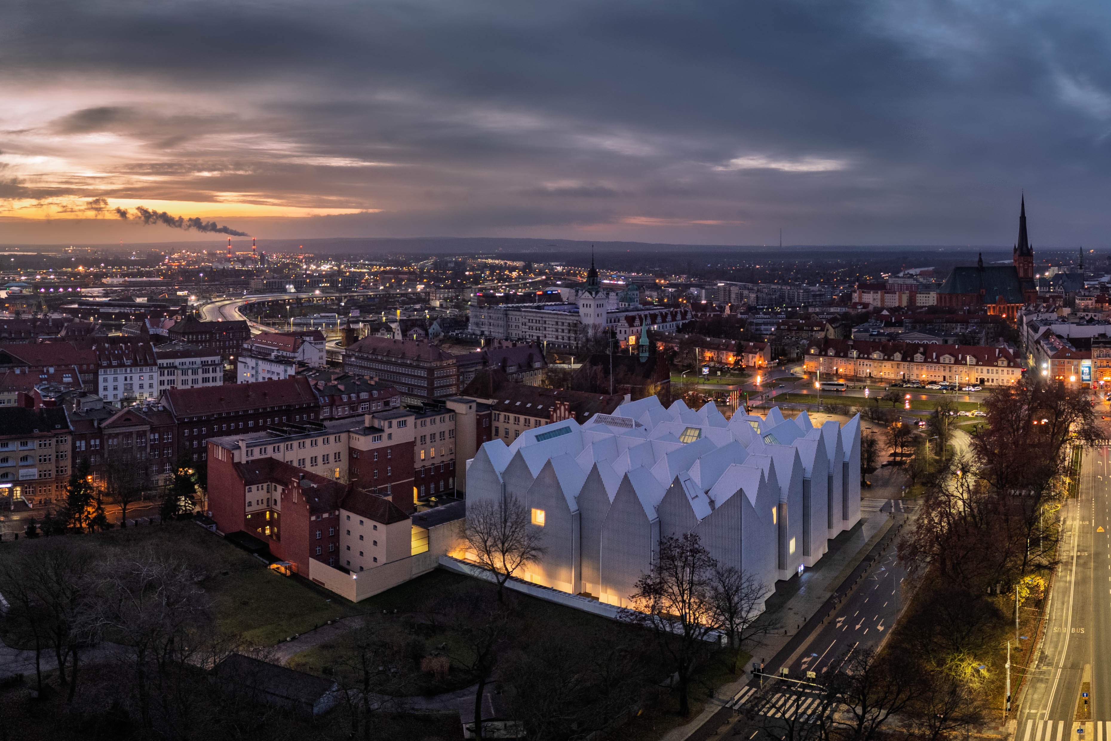 La Philharmonie de Szczecin vue du ciel au crépuscule, avec ses rues illuminées et la fumée qui s'élève à l'horizon.
