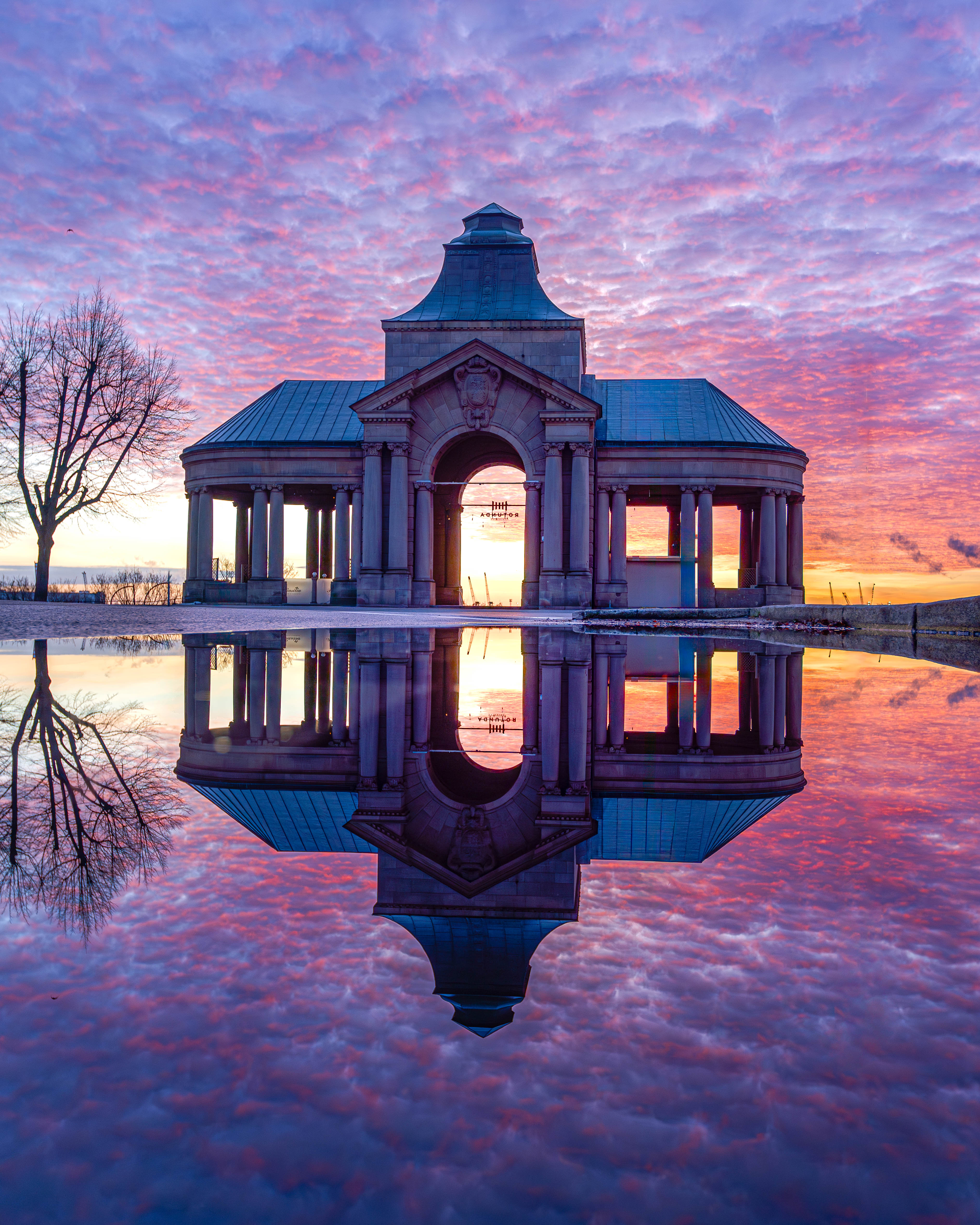 La rotonde située près des escaliers de Wały Chrobrego à Szczecin se reflète dans les eaux calmes, photographiée au coucher du soleil avec en toile de fond un ciel spectaculaire aux tons violets et roses.